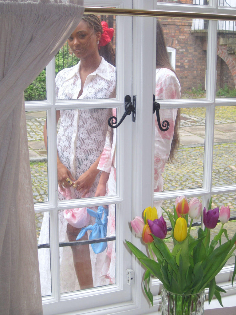 Woman standing behind a window with a vase of flowers in the foreground. They hold small knotted handbags and one wears a large pink scrunchie.